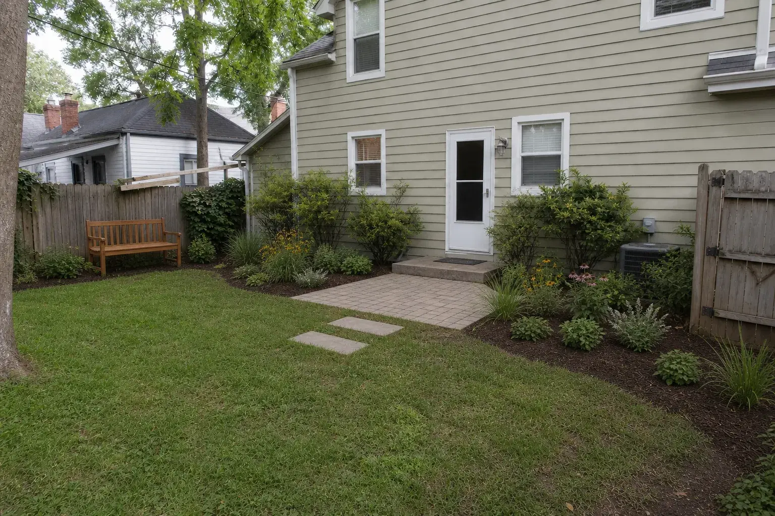 The same compact backyard with a modest paver patio, native planting beds, and cedar bench