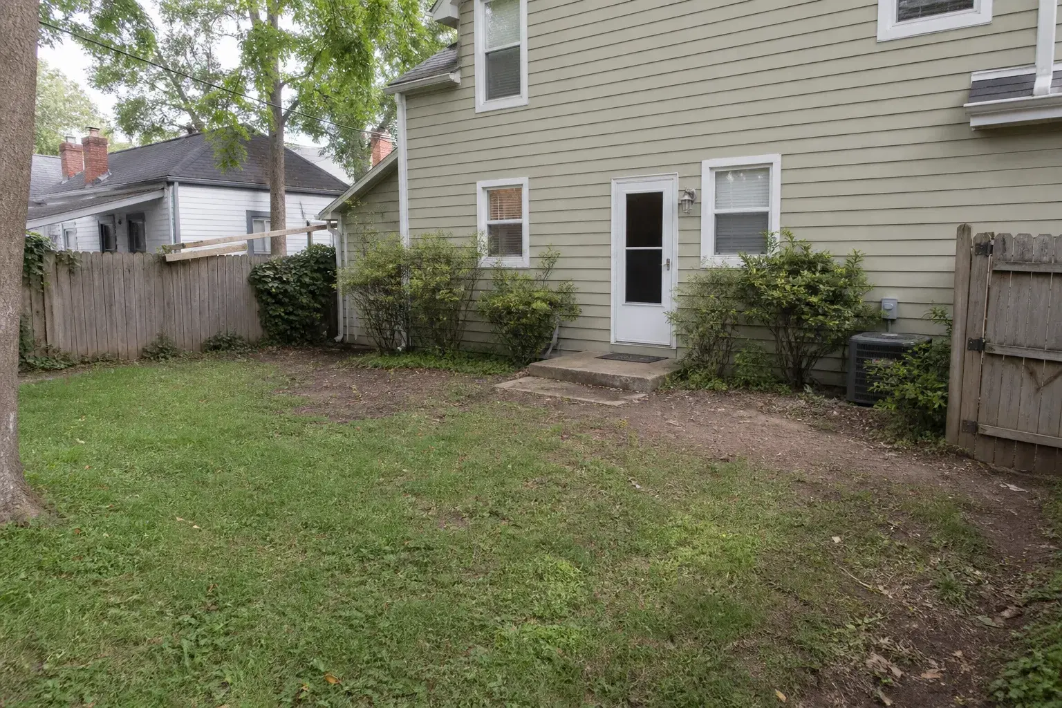 Compact backyard with patchy lawn, bare soil, worn concrete step, and sparse shrubs before landscaping